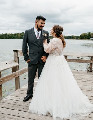A romantic wedding couple poses on a wooden dock by a lake, with the bride in a lace gown and groom in charcoal suit.