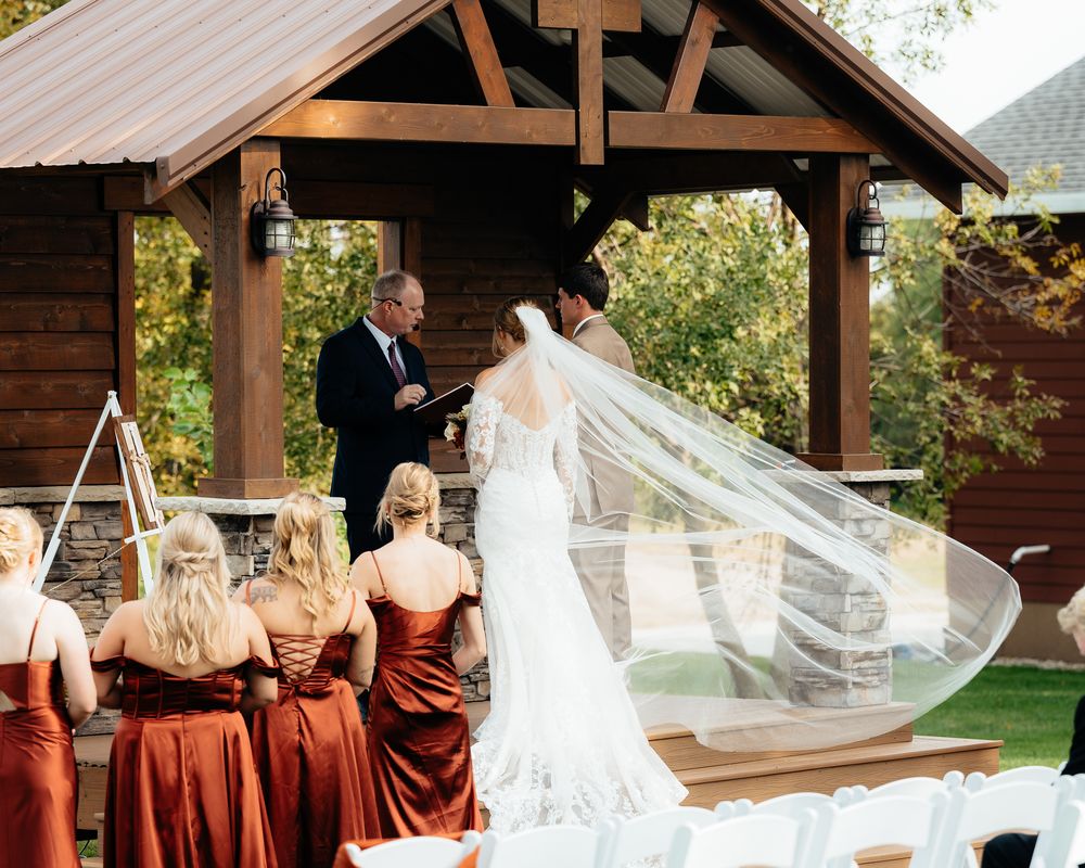 An outdoor wedding ceremony under a wooden pavilion with bridesmaids in burgundy dresses and a flowing white veil catching the breeze.