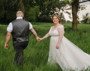 A couple in wedding attire walks hand in hand through a lush green field during golden hour.