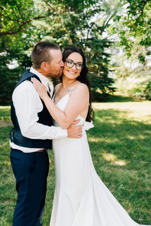 A joyful wedding couple embraces in a sunlit garden setting with lush greenery in the background.