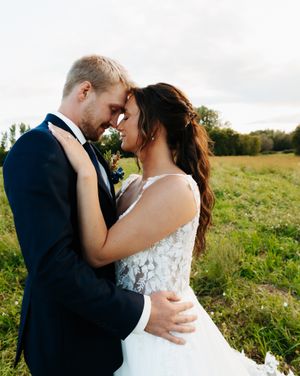 Newlyweds share an intimate embrace in a sunlit meadow with wildflowers and rolling hills in the background.