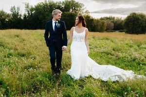 A wedding couple stands together in a golden meadow at sunset with the bride's dress trailing behind.
