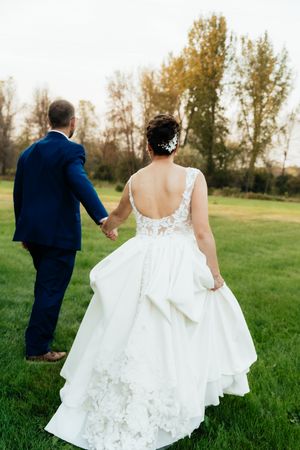 A couple walks hand in hand across a grassy field, with the bride wearing a flowing white lace gown with open back detail.