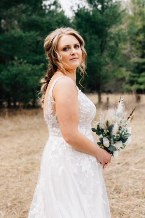 A bride in a lace wedding gown holds a white bouquet while posing in a rustic outdoor setting.