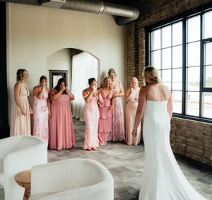 Wedding party in pink bridesmaid dresses gathered in an industrial brick venue with large windows.