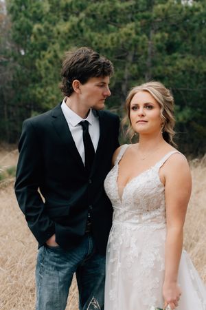 A bride in a lace wedding dress stands with her casual-formal dressed partner against a backdrop of evergreen trees.