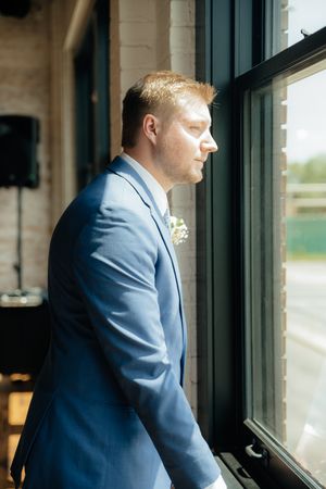 A person in a light blue suit gazes contemplatively out of a large window in an indoor setting.