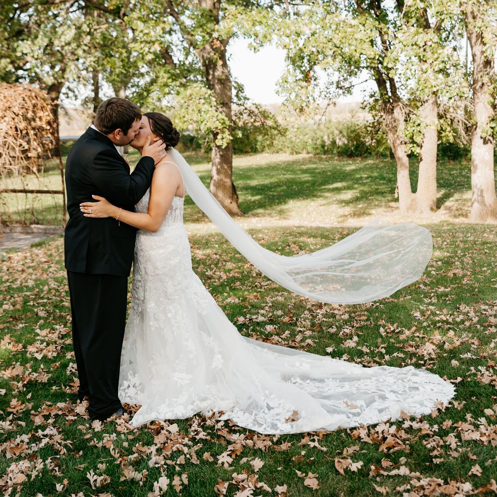 A long flowing wedding veil catches the autumn breeze in a scenic outdoor setting with fall foliage on the ground.