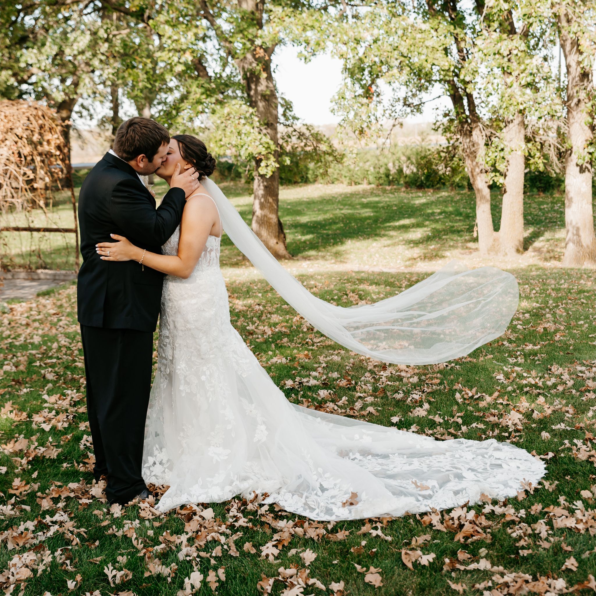 A long flowing wedding veil catches the autumn breeze in a scenic outdoor setting with fall foliage on the ground.