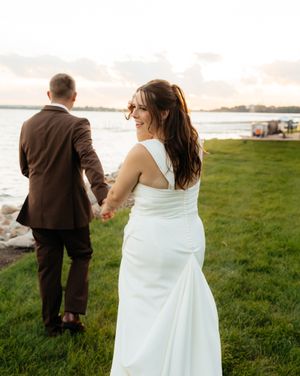 A newlywed couple walks hand in hand along a waterfront path during golden hour.