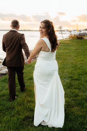 Couple walks hand in hand across a grassy field at sunset near the water.