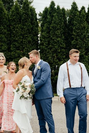 Wedding party poses together outdoors with bridesmaids in coral dresses and groomsmen in navy suits with suspenders.