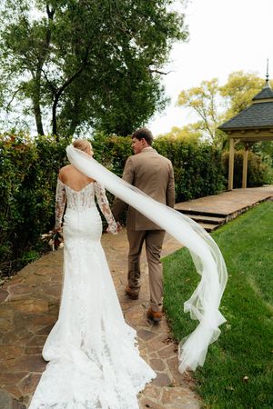 A flowing white wedding veil catches the breeze on a stone pathway surrounded by greenery.
