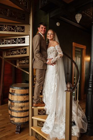 A couple poses on rustic wooden stairs next to a vintage barrel in an elegant wedding setting.