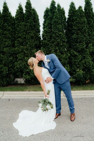 A romantic wedding couple shares a dip kiss pose in front of a row of evergreen trees.