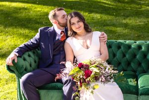 Romantic couple poses on emerald green tufted sofa with vibrant floral bouquet outdoors.