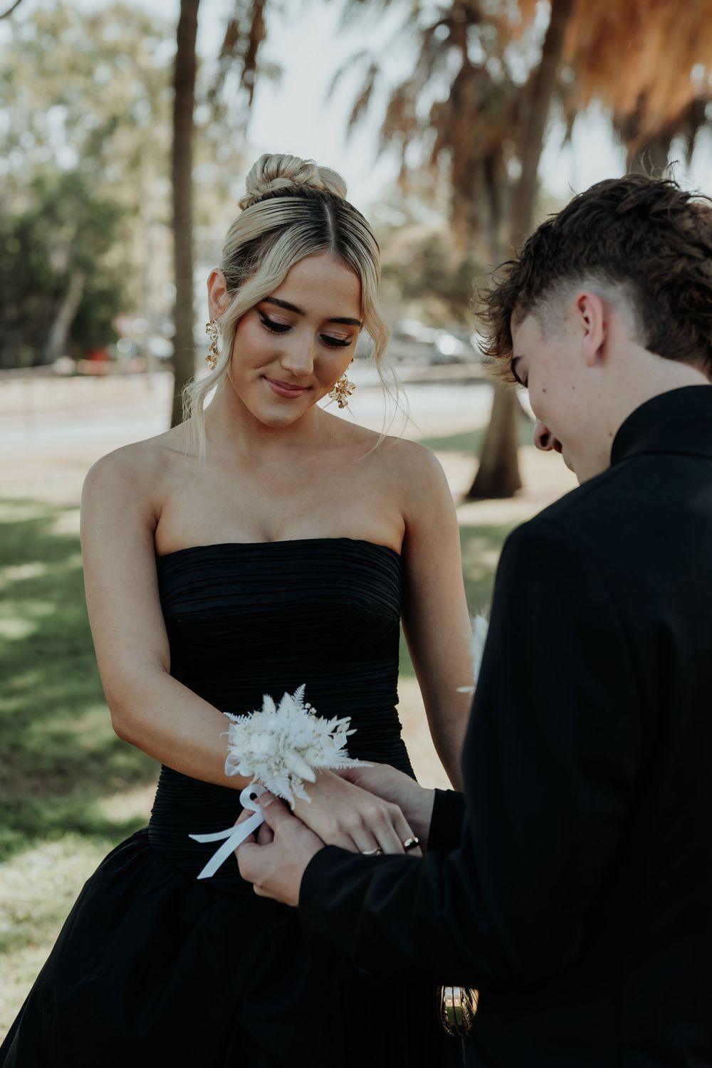 Formal couple in black attire exchanging corsage in outdoor gladstone garden setting.
