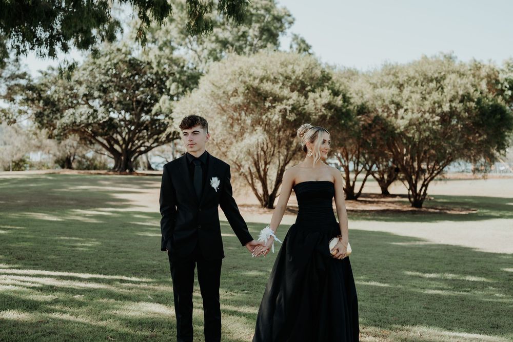 Elegant couple in formal black attire walking through sunlit gladstone garden path.