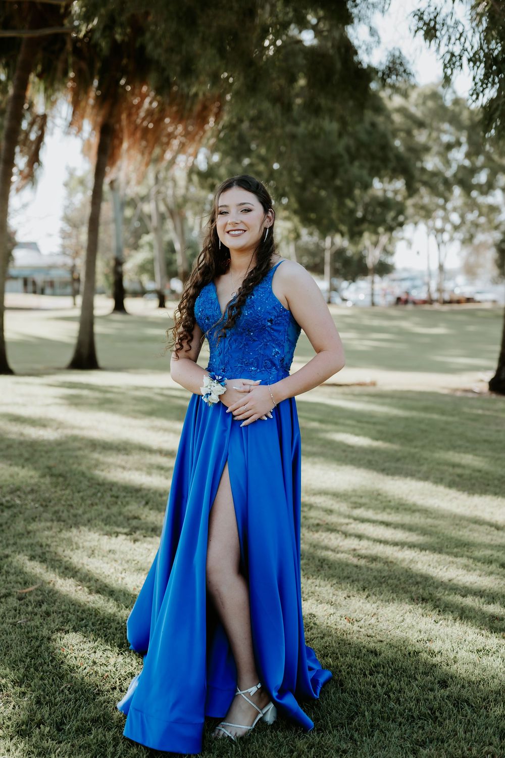 A flowing royal blue formal dress photographed in a sunlit park setting with trees in the background.