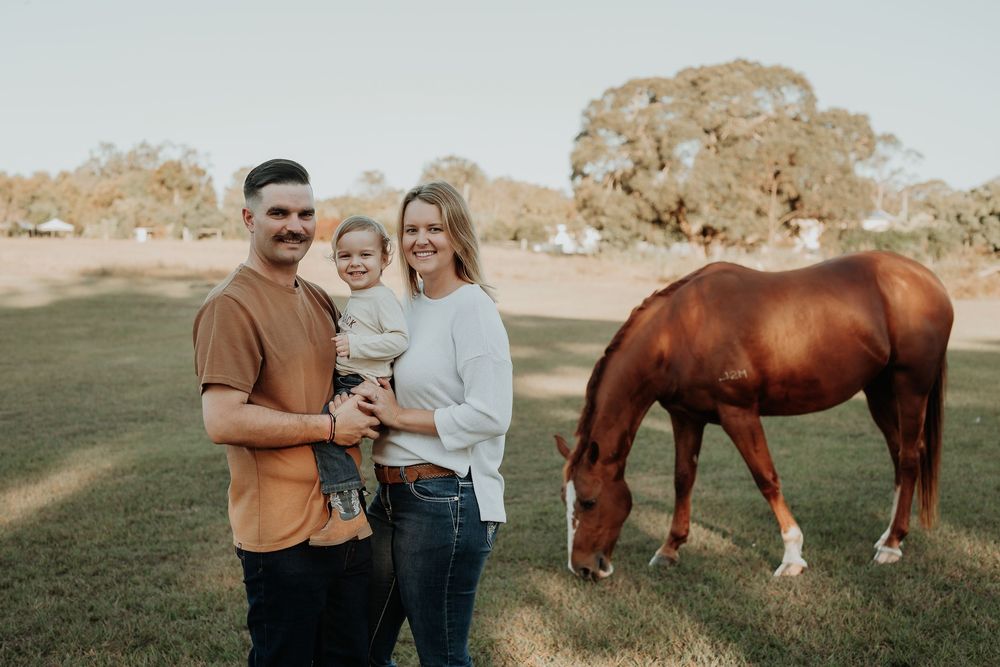 A smiling family with a baby poses beside a grazing chestnut horse in a sunny green paddock.