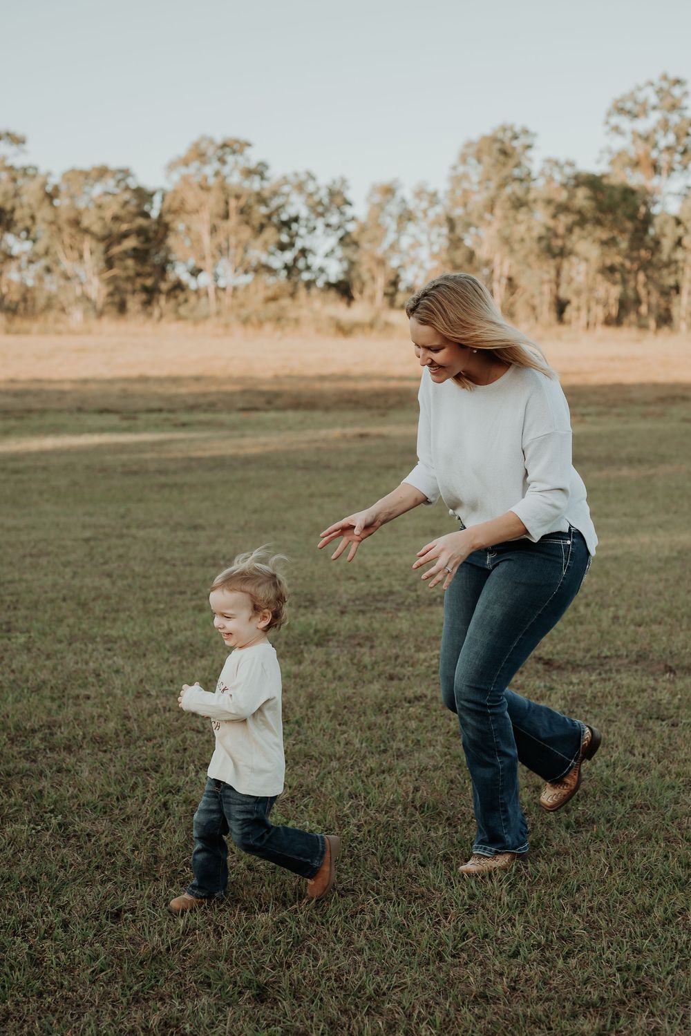 A mother and toddler playing together in a grassy field, both wearing white tops and jeans on a sunny day.