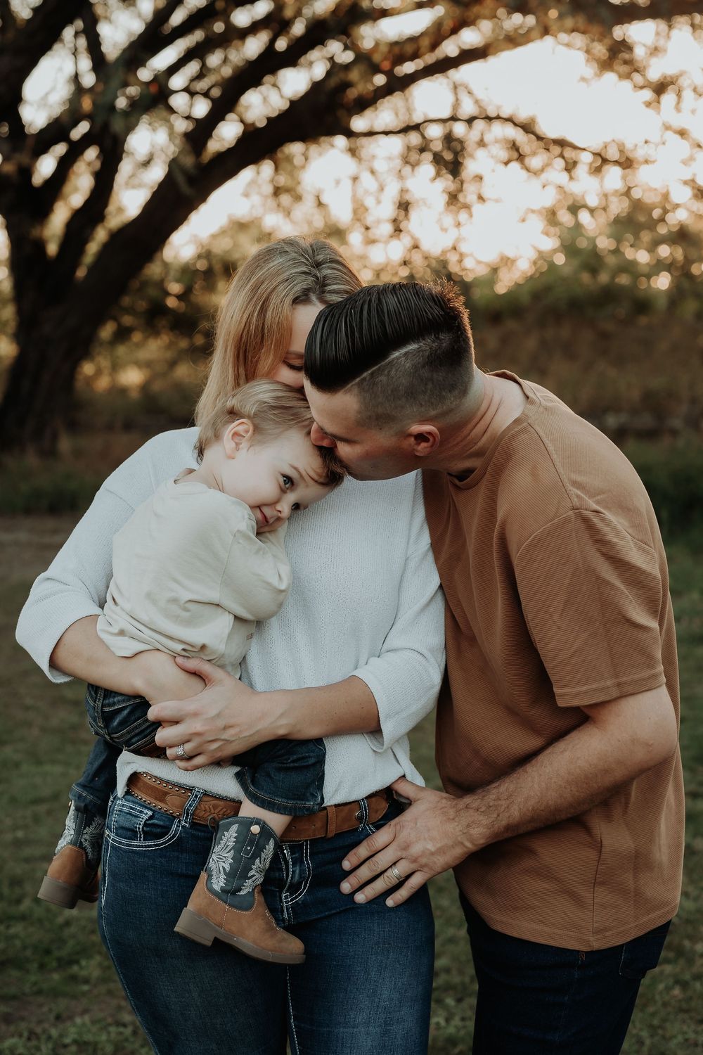 A loving family of three shares a tender moment outdoors, with parents embracing their young child at golden hour.