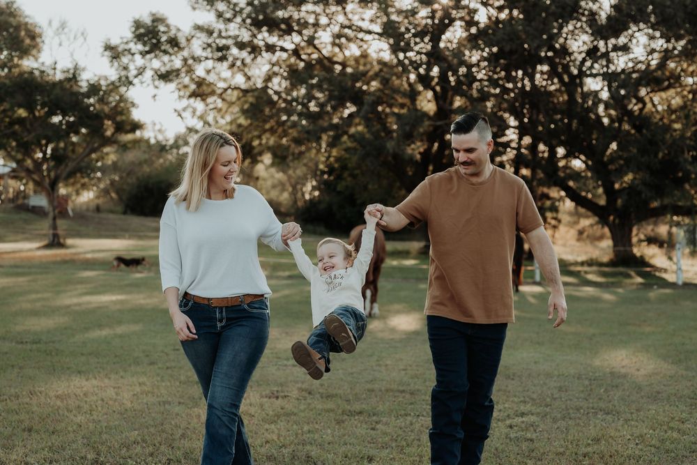 A happy family swings their young child between them while walking in a sunny green park.