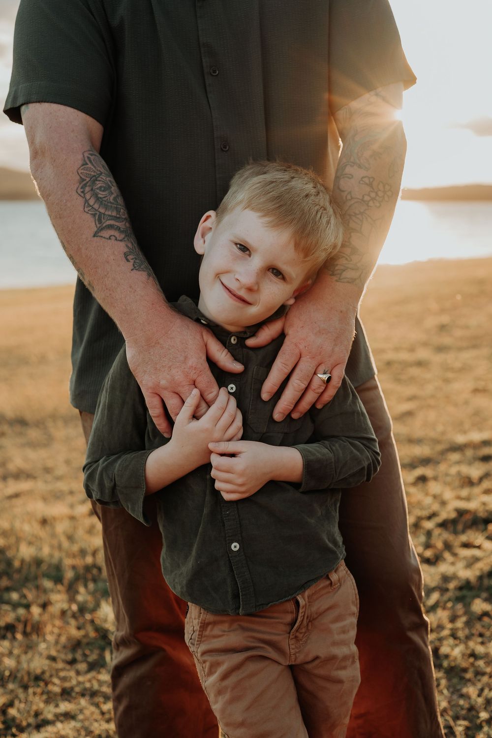 A tattooed father hugs his smiling young blond son outdoors at golden hour near a lake.