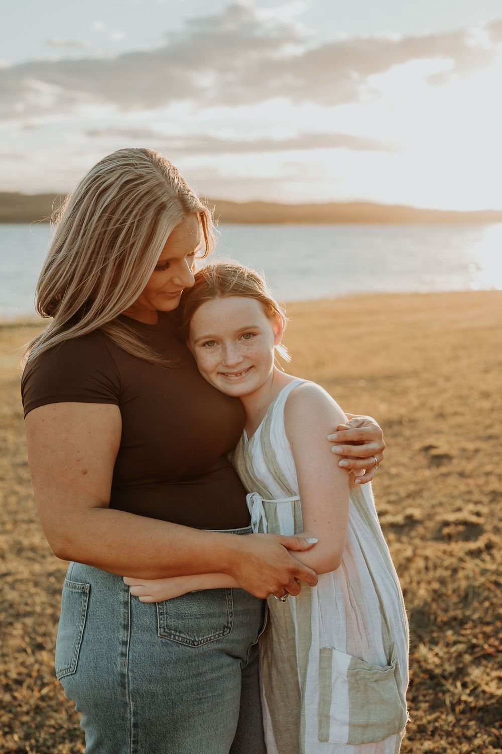 A mother and daughter share a warm embrace on a golden beach at sunset, with calm water and clouds in the background.