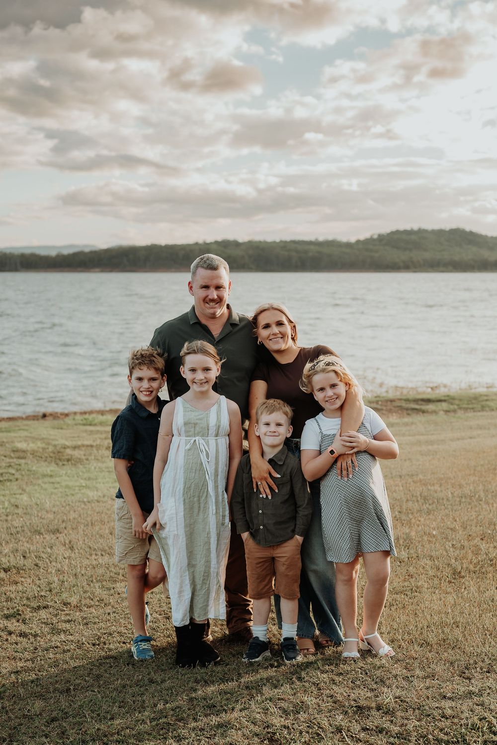 Family of six posing outdoors near a lake at sunset, parents with four young children smiling on green grass.