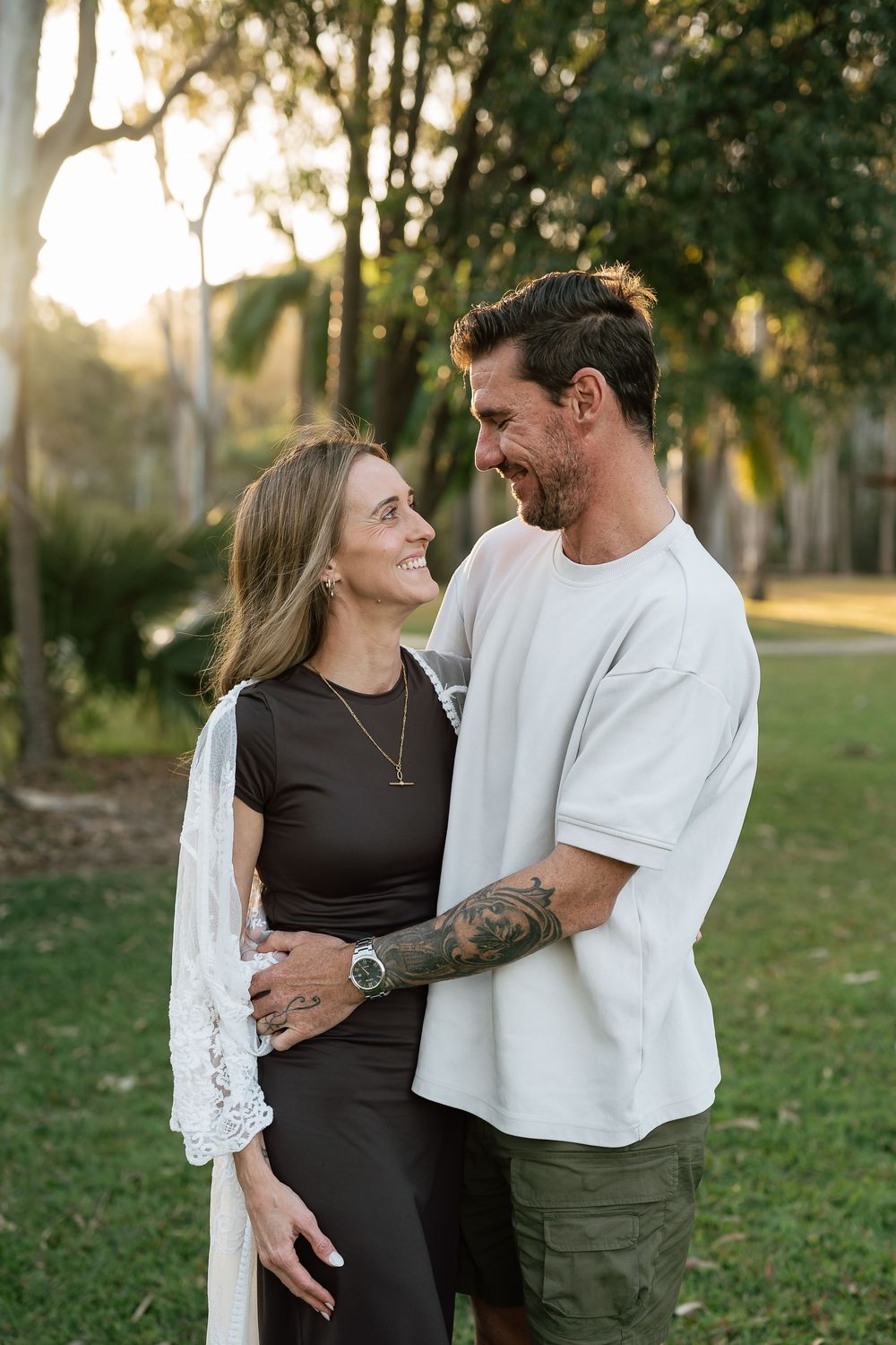 A smiling couple embraces outdoors at golden hour, with lush green trees in the background, the man wearing a white tee.
