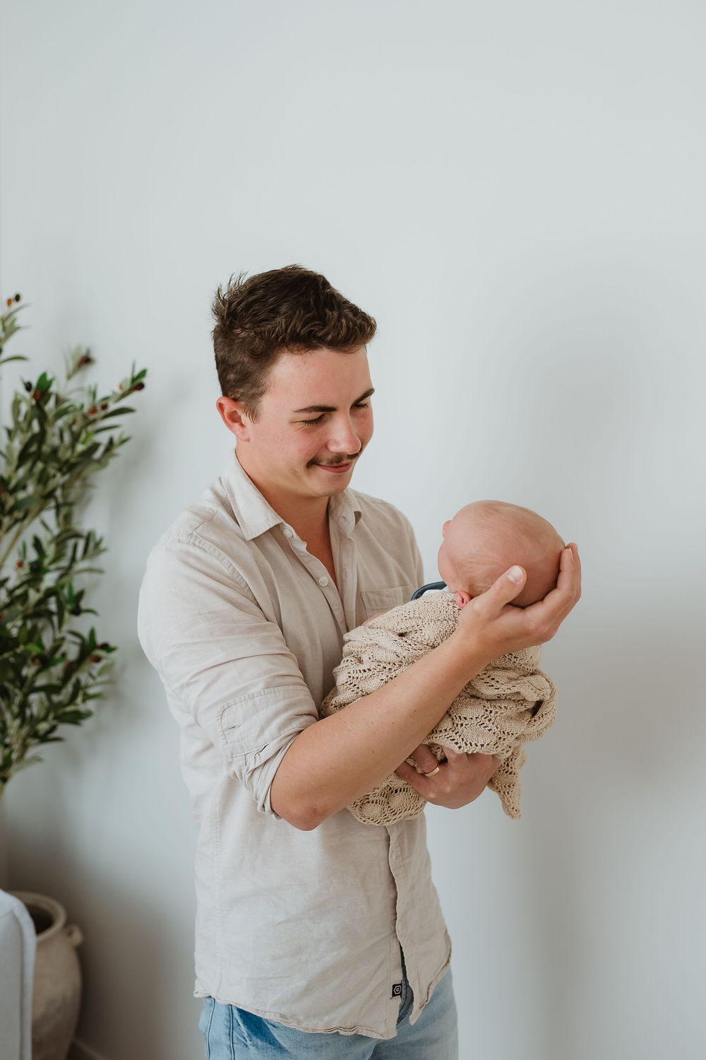 Young father with mustache tenderly holds newborn baby wrapped in cream knit blanket against white wall.