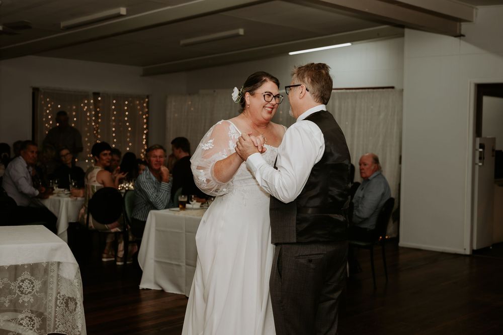 Bride and groom share their first dance at wedding reception with guests watching in background.
