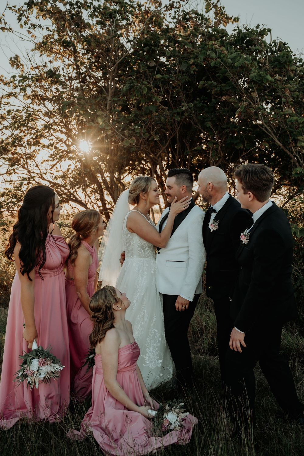 Gladstone wedding party in pink bridesmaid dresses and black suits gathered under a tree at sunset.