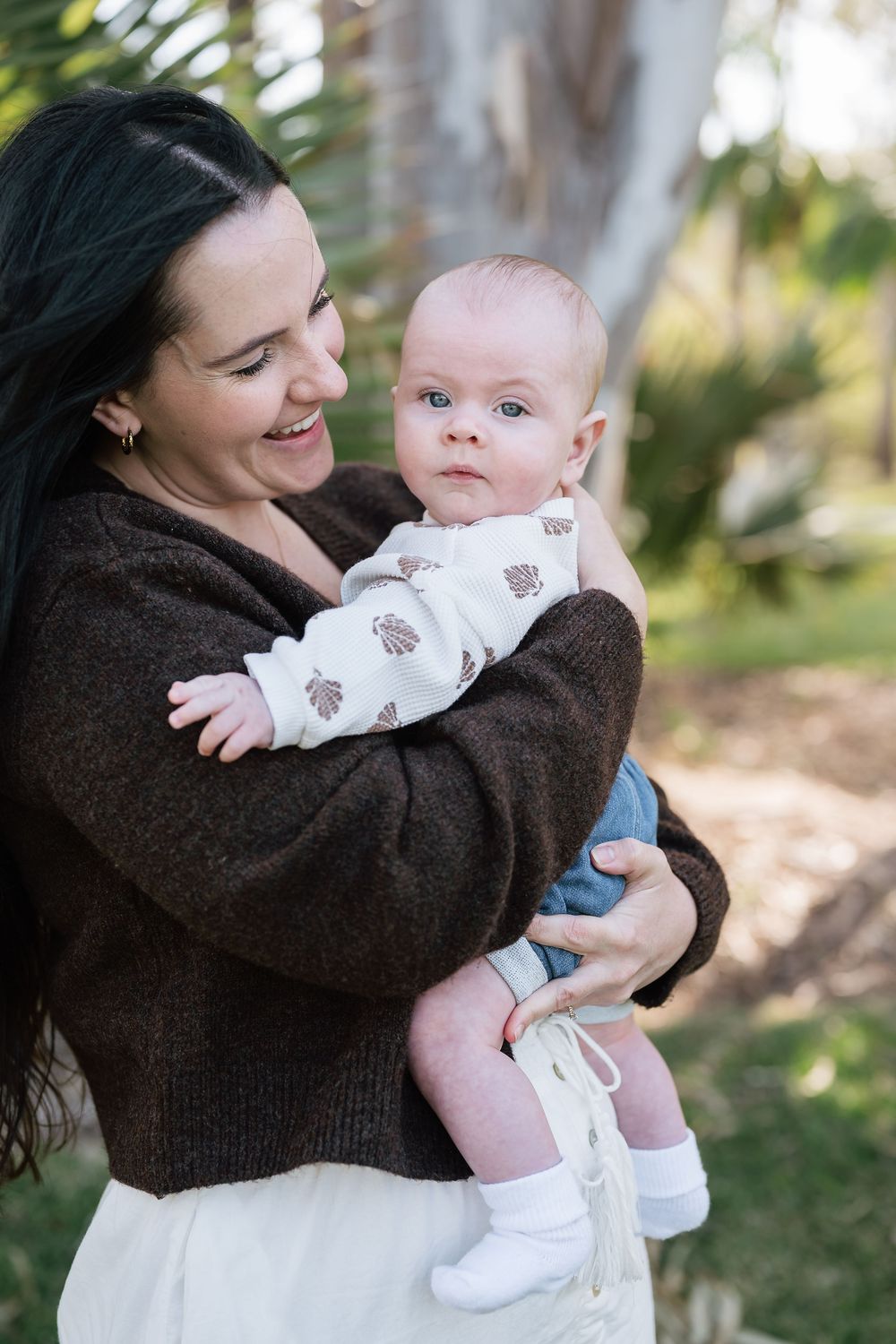 A smiling dark-haired woman in a brown sweater holds an alert baby outdoors near palm trees.