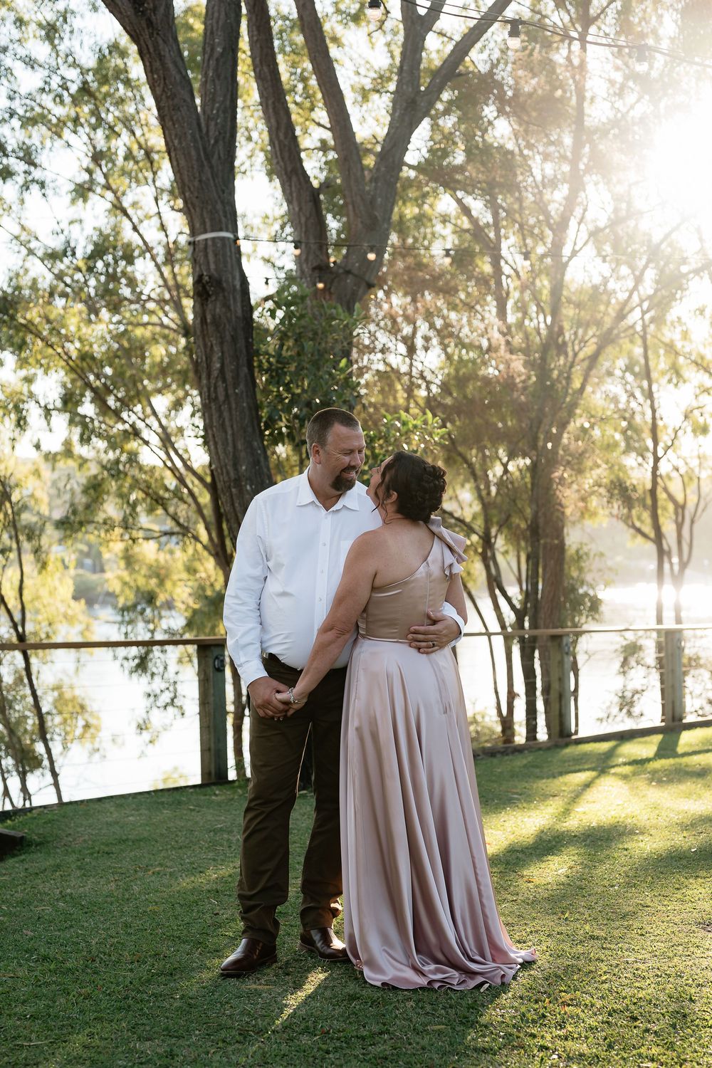 Couple sharing a romantic moment on a sunlit lawn by a river, man in white shirt and woman in dusty pink gown.