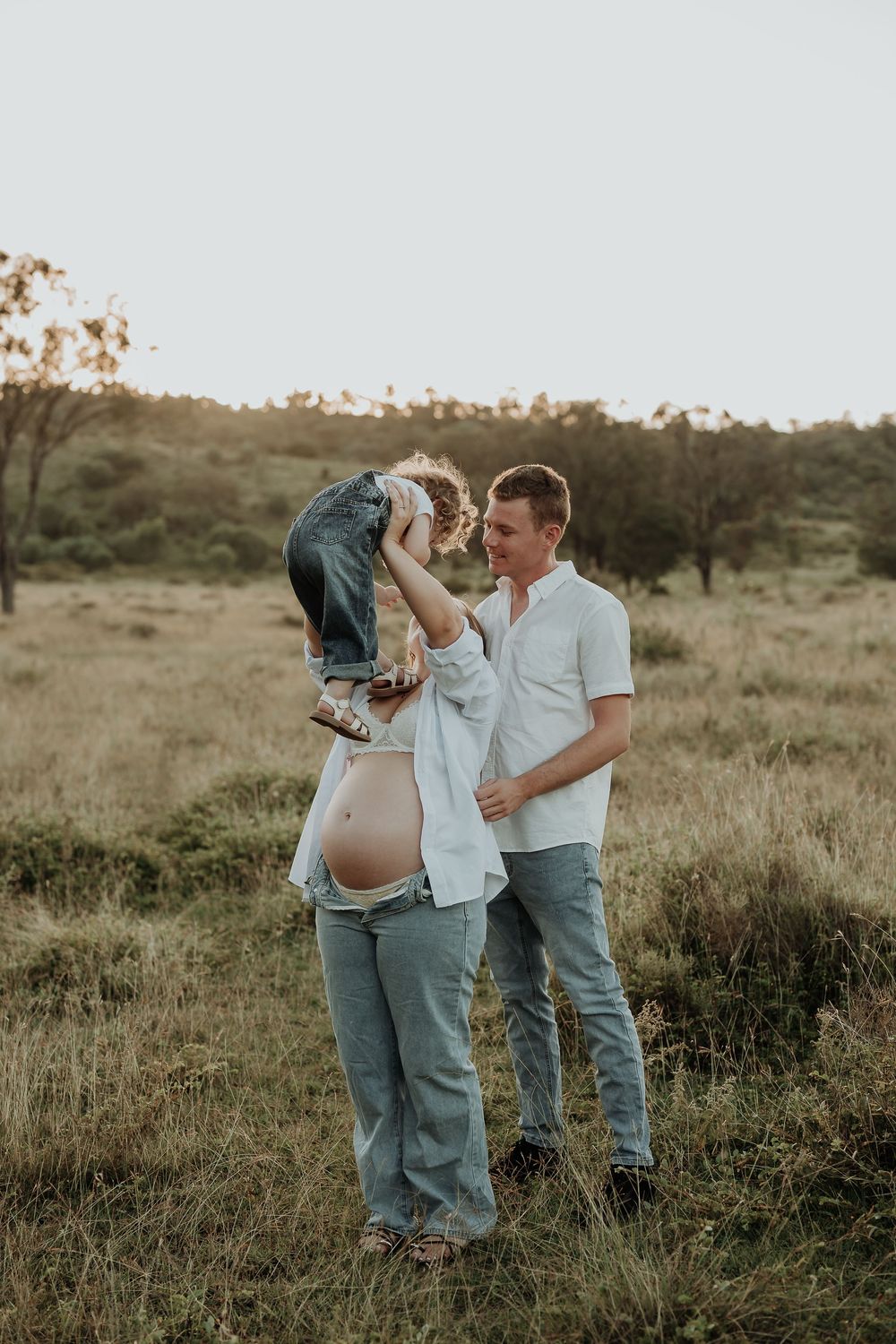 Pregnant woman and man playing with toddler in golden hour field, maternity family photo outdoors.