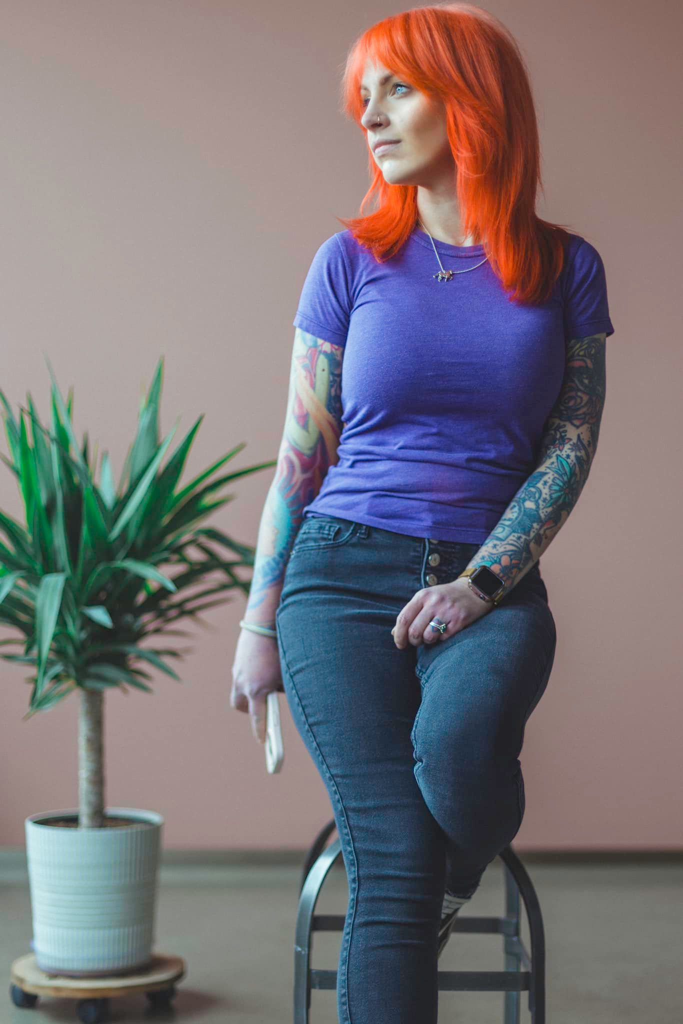 Person in blue shirt with colorful tattoos sitting near a potted plant.