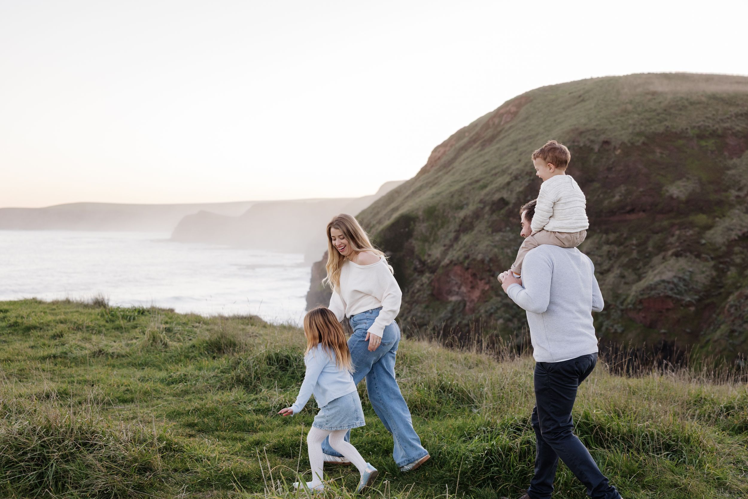 Family of four walking on coastal cliffside grass with ocean views at sunset.