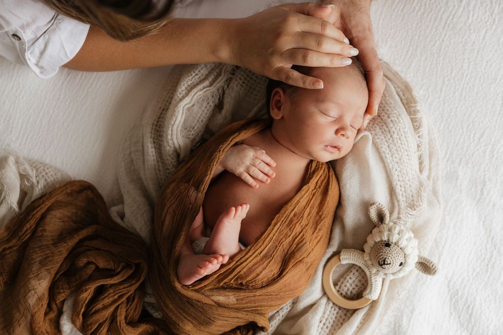 Sleeping newborn baby wrapped in brown swaddle on white blanket with wooden teething toys.