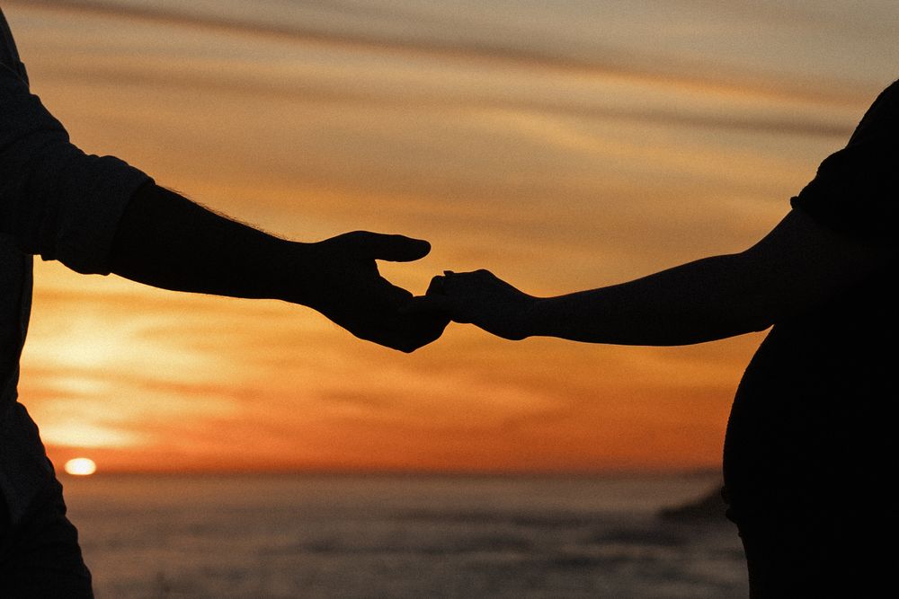 Silhouetted hands reach across dramatic orange sunset sky creating romantic connection.