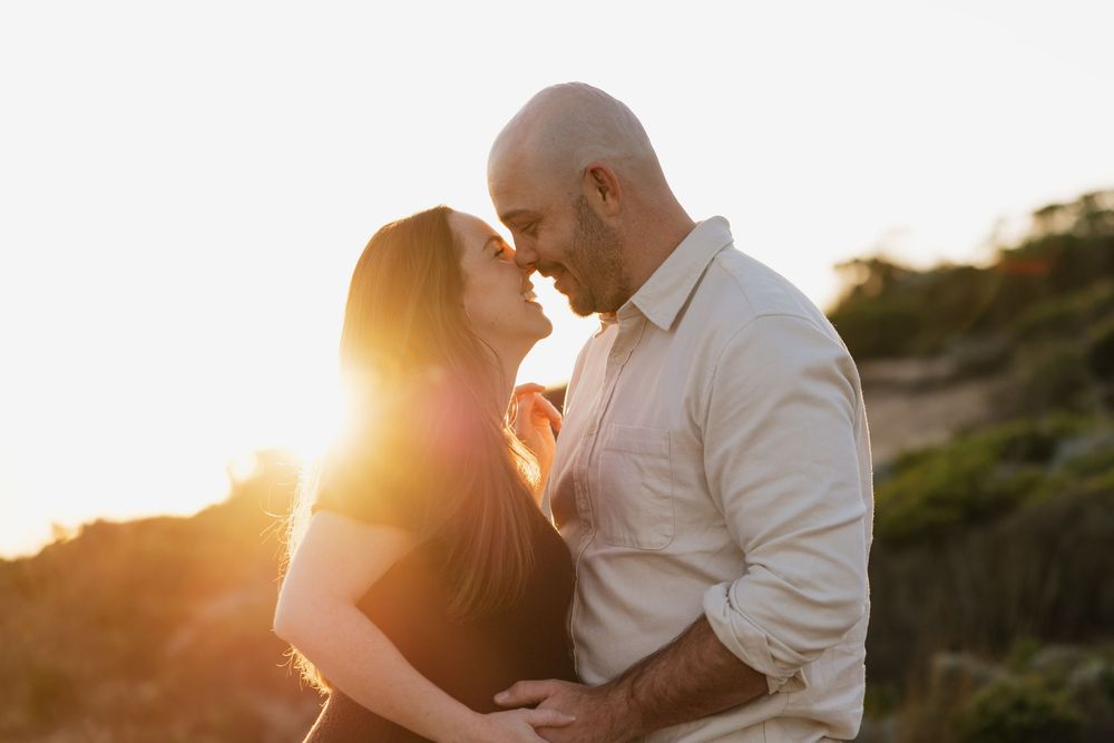 Couple sharing romantic moment at sunset along ocean cliffs in white summer outfits.