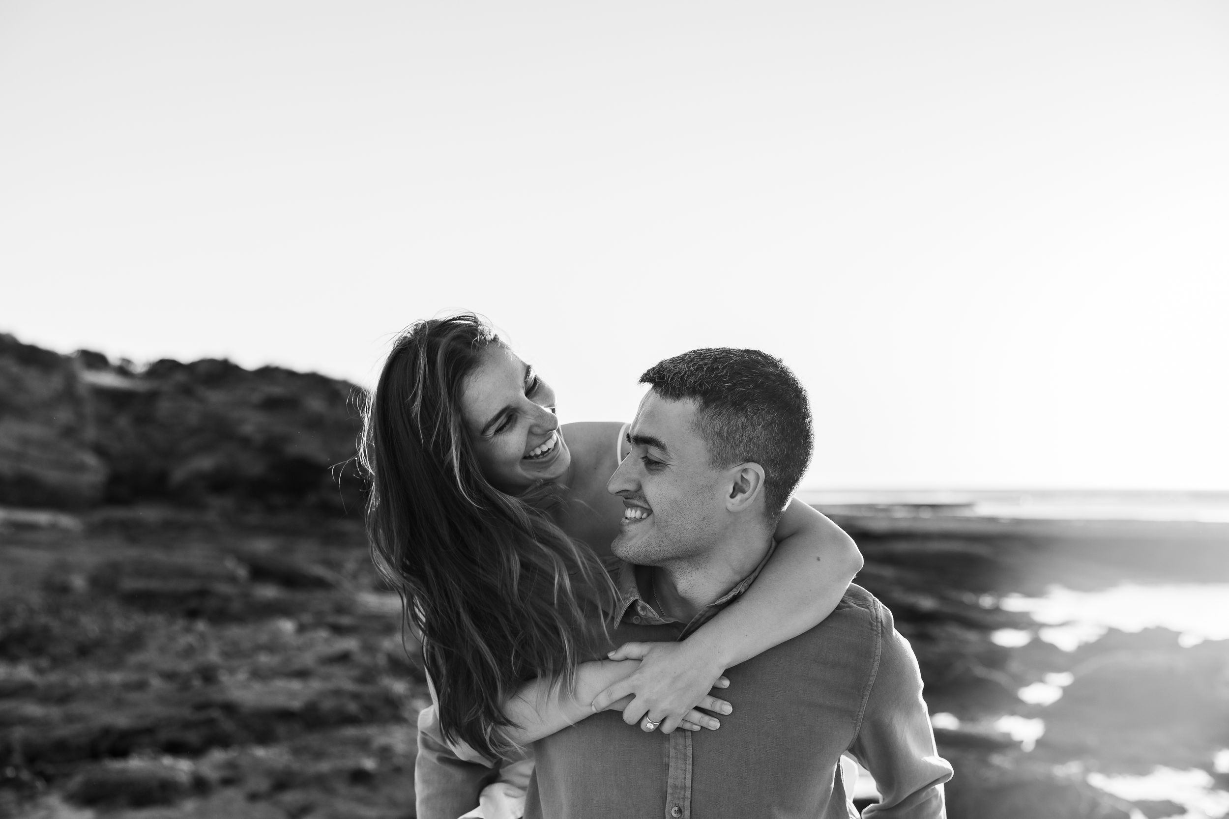 A joyful couple sharing an intimate black and white moment together on a scenic rocky coastline at sunset.