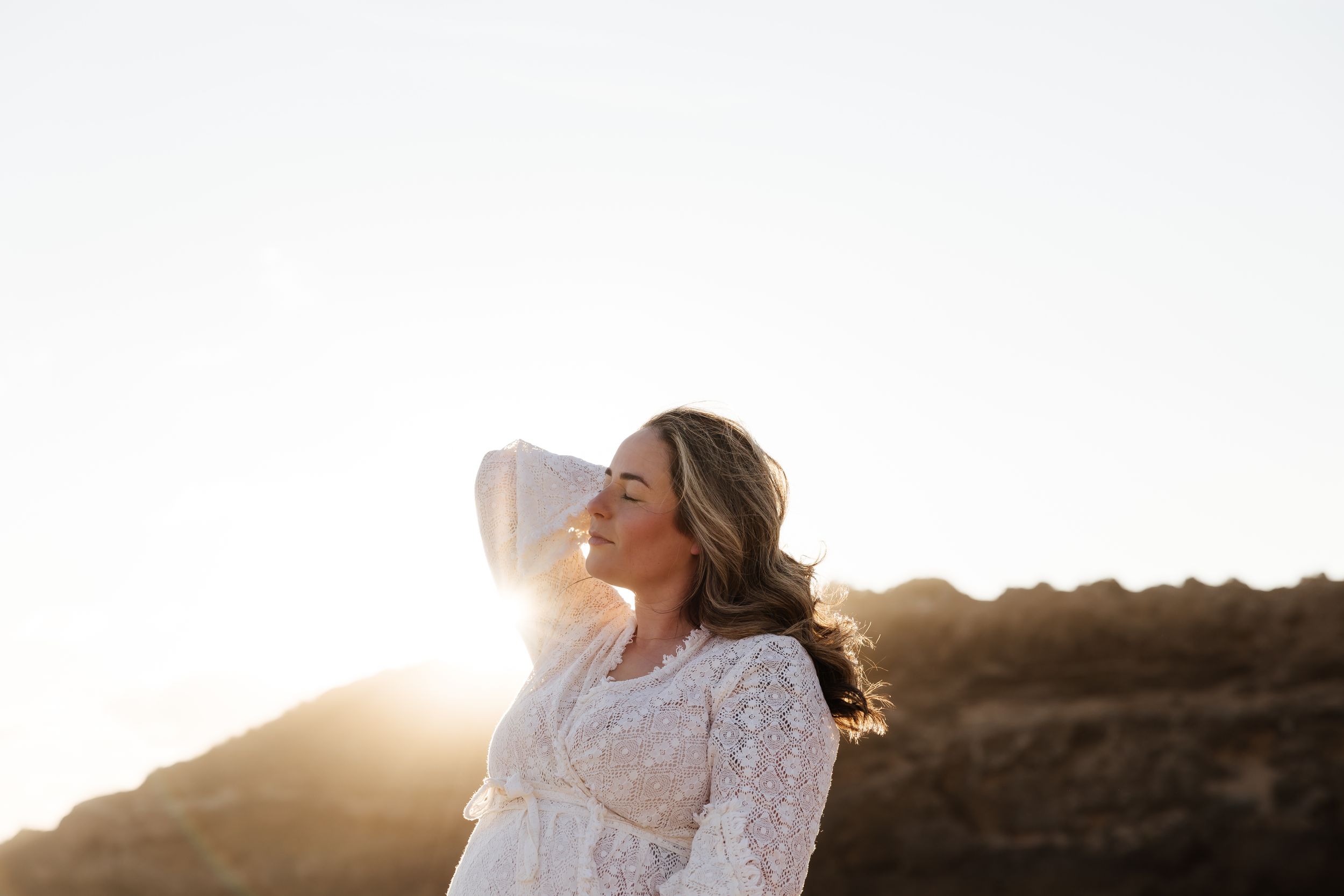Woman in white lace dress standing in golden sunset light against mountainous backdrop.
