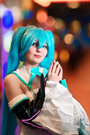 Woman cosplaying as Hatsune Miku with teal twin-tail wig and white dress with hands pressed together.