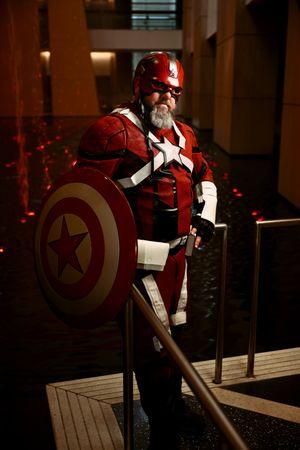 Man with beard cosplaying as Red Guardian in red Captain America-style armor holding shield.