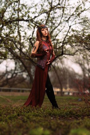 Woman in full Scarlet Witch red costume with crown standing among blooming trees outdoors.