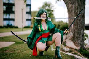 Woman in green cosplay costume crouching with two swords held in her mouth outdoors.