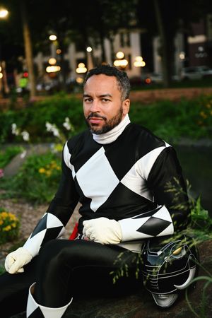 Man in black and white Power Ranger costume sitting outdoors at night with helmet beside him.