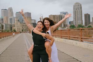 Two women celebrating on a bridge, one giving the other a piggyback ride with city skyline behind.